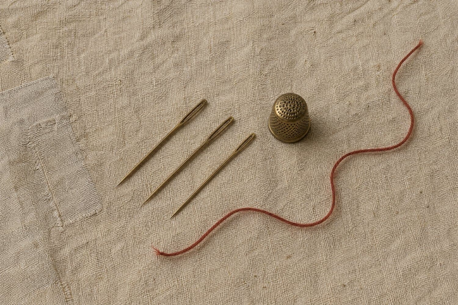 Three brass darning needles and a small thimble laid in a diagonal on unbleached linen with a length of madder thread beside them