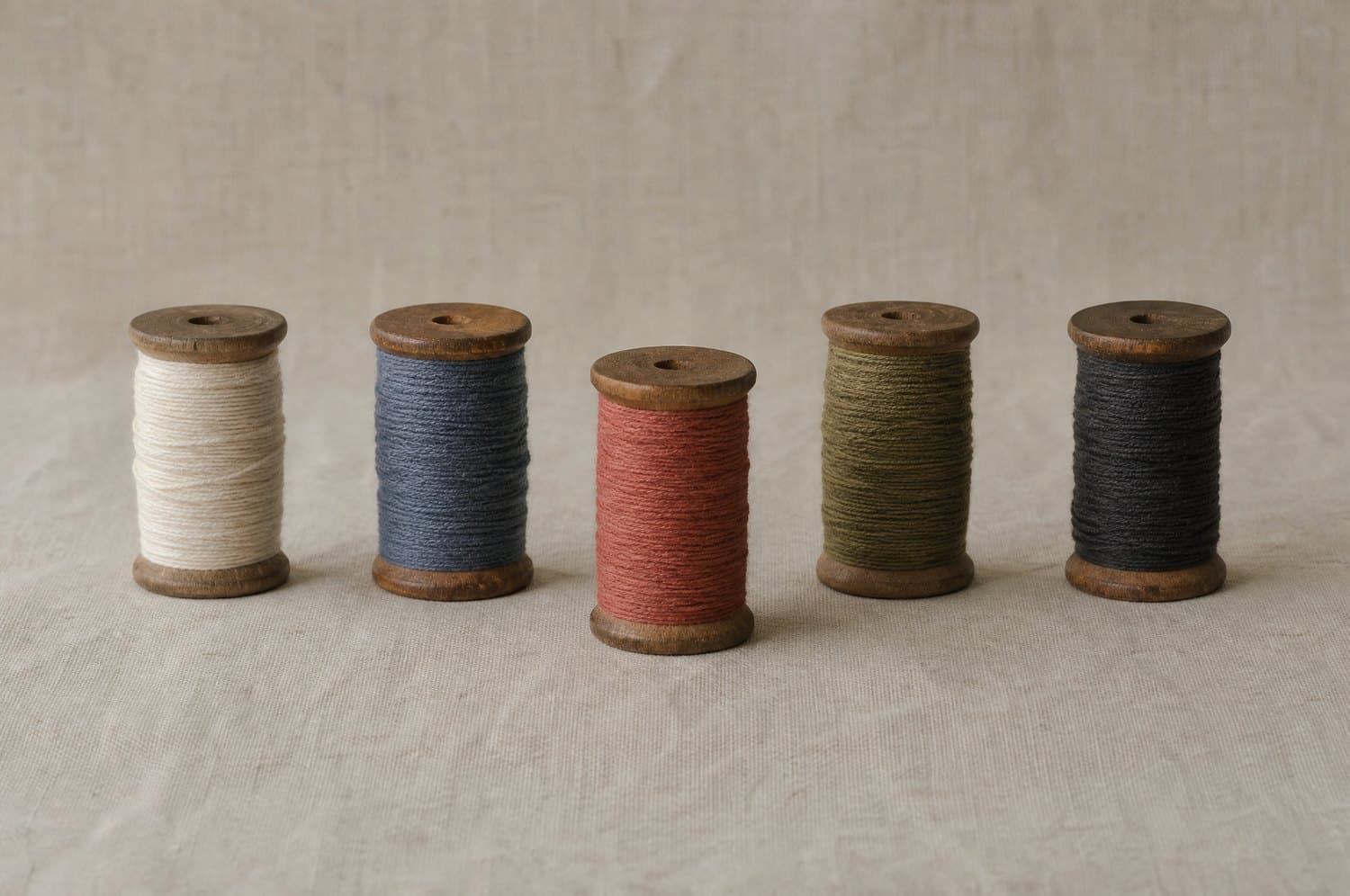A row of wooden thread spools in cream, indigo, madder, olive and charcoal standing on unbleached linen
